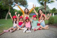 Joyful children and two women sitting together in a park, raising hands and smiling.